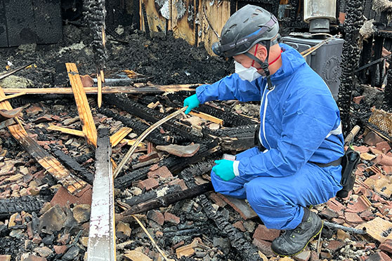 Fire investigator in protective gear inspecting fire-damaged debris at a burned site.